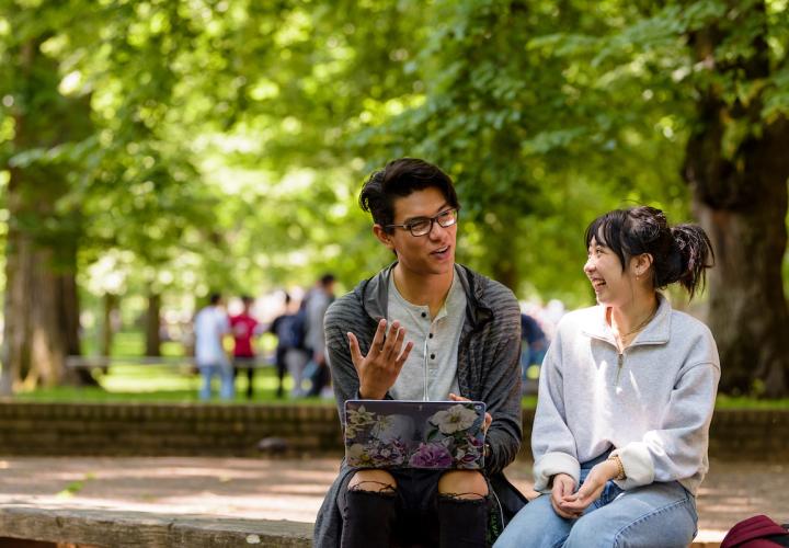 One male and one female student sit outside sitting next to each other. The male student has his laptop in his lap and is talking to the female student.