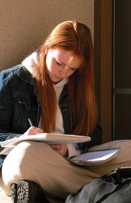 Photo of a student sitting against a wall writing notes in her booklet